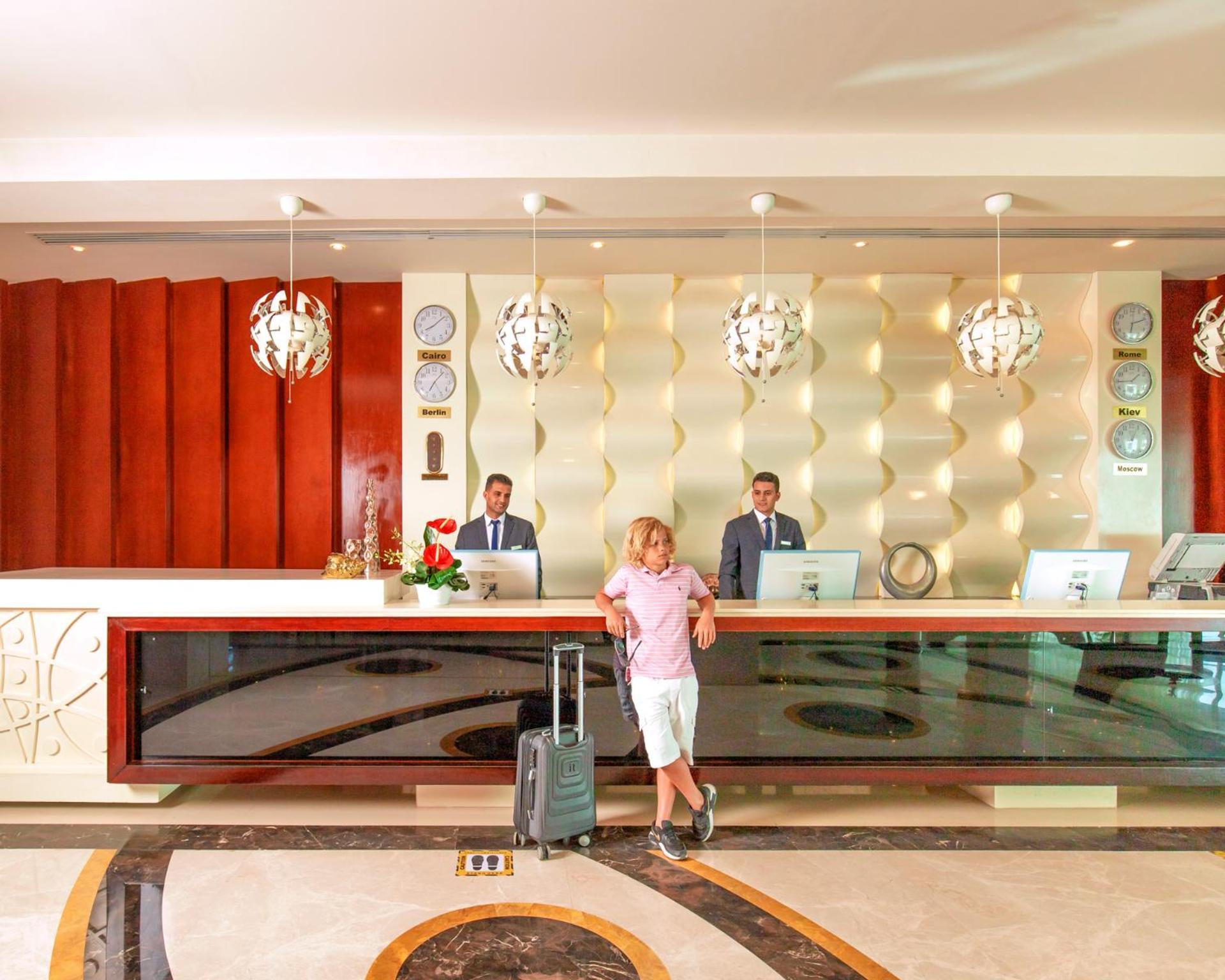 Hotel Royal Albatros Moderna - Families and couples only à Charm el-Cheikh Extérieur photo The photo depicts a hotel reception area. At the front desk, two staff members in suits are standing, presumably ready to assist guests. A man in casual attire, holding a suitcase, is standing in front of the desk. The reception has a modern design w