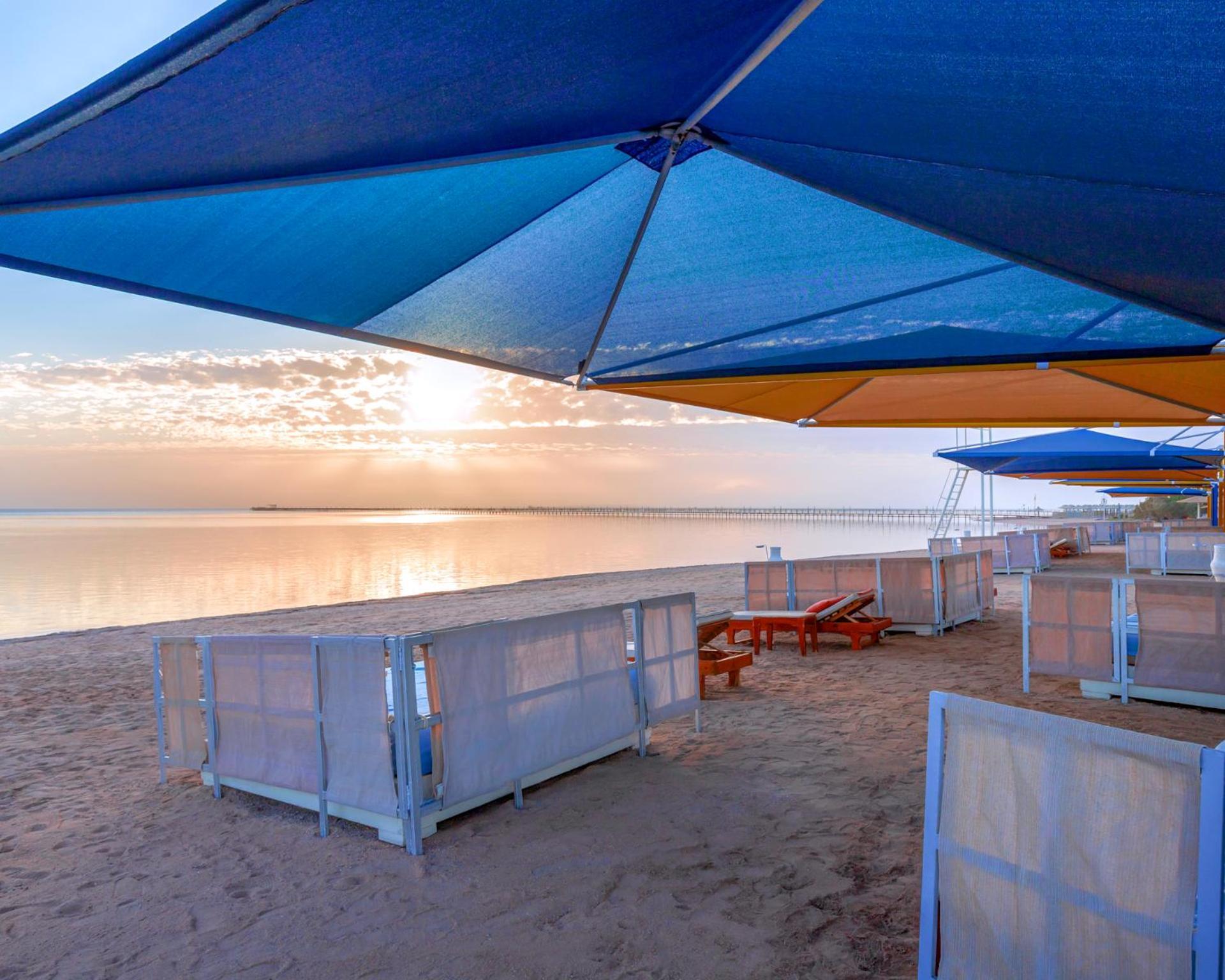 Hotel Royal Albatros Moderna - Families and couples only à Charm el-Cheikh Extérieur photo The photo shows a beach scene during sunset. There are colorful umbrellas in various shades of blue and orange set up over sandy areas. In the foreground, there are some wooden lounge chairs, and behind them, partition-like structures or beach cabana