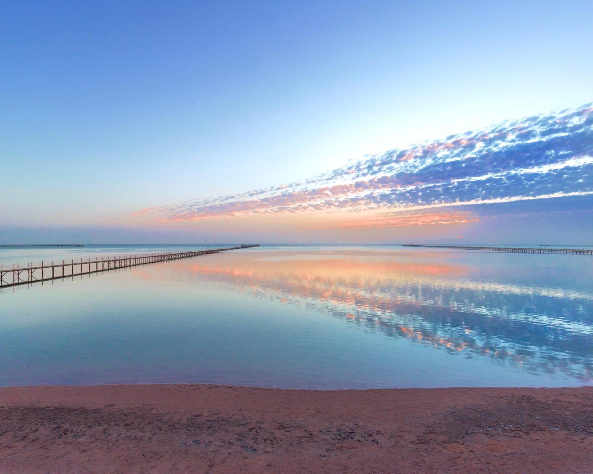 Hotel Royal Albatros Moderna - Families and couples only à Charm el-Cheikh Extérieur photo The photo depicts a serene coastal scene at dawn or dusk. There is a calm body of water that reflects the soft hues of the sky, which include shades of blue, pink, and orange. A long wooden pier extends into the water, adding depth to the composition