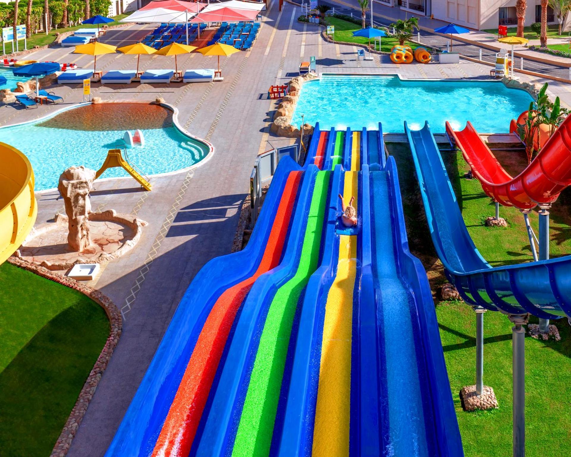 Hotel Royal Albatros Moderna - Families and couples only à Charm el-Cheikh Extérieur photo The photo shows a vibrant water park featuring several large water slides in various colors, including red, blue, green, and yellow. There are swimming pools nearby, one of which has a clear blue appearance. The area is surrounded by green grass and 